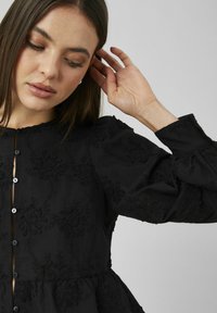 Robe noire à manches longues avec broderie florale, encolure ronde et boutons sur le devant ; tissu doux avec une texture apparente.