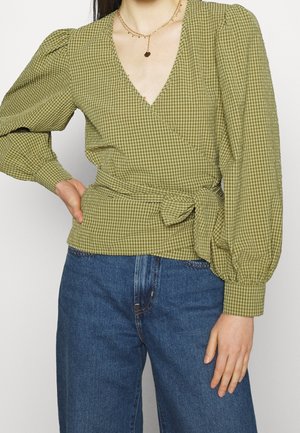 Frau trägt grüne karierte Wickelbluse mit Puffärmeln und blaue Jeans, accessoriert mit geschichteten goldenen Halsketten.