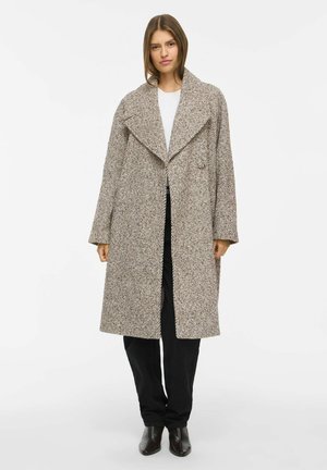 Femme portant un long manteau beige texturé par-dessus un haut blanc, un pantalon noir et des bottines noires, debout devant un fond blanc uni.