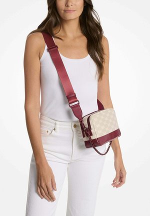 Femme portant un débardeur blanc côtelé et un pantalon, portant un sac bandoulière crème et bordeaux avec une large sangle réglable et un détail chaîne.