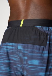 Men's swim shorts feature a multicolor blue and black design with a black waistband and a black mesh pocket on the back. Neon accent detail.