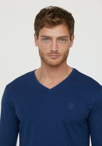 Chemise à manches longues bleu marine en mélange de coton avec un col en V et un petit logo circulaire sur la poitrine gauche. Texture lisse, design ajusté.