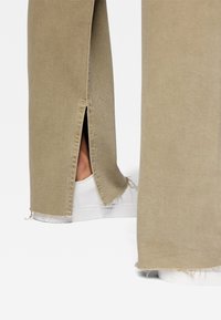 Pantaloni beige a gamba larga con spacco laterale e orlo sfilacciato indossati con sneakers bianche su uno sfondo bianco.