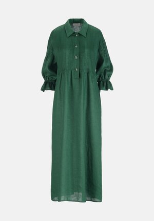 Frau trägt ein langes, grünes, knöpfbares Kleid mit Puffärmeln, hält eine beigefarbene gewebte Clutch und trägt Goldschmuck sowie Sandalen.