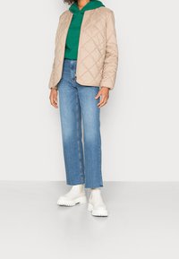 Beige doorgestikte jas met een rits aan de voorkant, groene capuchontrui eronder, lichtblauwe jeans met rechte pijpen en witte laarzen met dikke zolen.