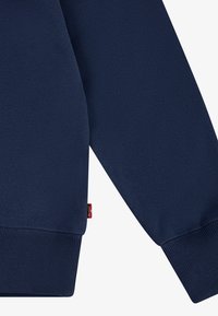 Námornícka modrá mikina s rebrovaným spodným okrajom a manžetami, mäkkou textúrou a malou červenou logovou nálepkou na boku. Jednoduchý, klasický dizajn.