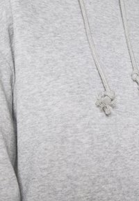 Felpa grigia con tessuto testurizzato, dotata di cappuccio e lacci con estremità annodate. Il design casual enfatizza il comfort e la semplicità.