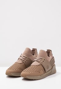 Par de zapatos deportivos de tono beige con parte superior perforada, banda elástica ancha y suelas de goma texturizadas sobre superficie blanca.