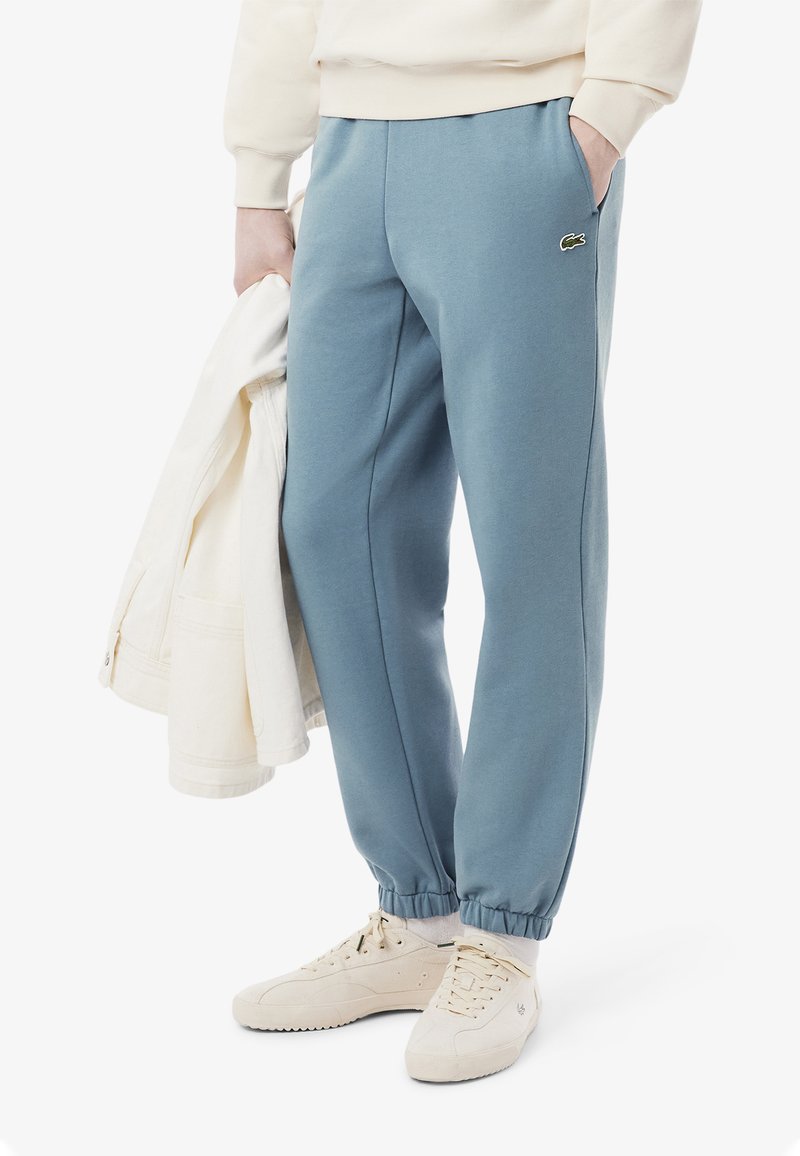 Vaaleansiniset collegehousut, jotka on valmistettu puuvillasekoitteesta. Housuissa on sivutaskut, elastiset hihnat ja pieni vihreä Lacosten logo vasemmalla etupuolella.