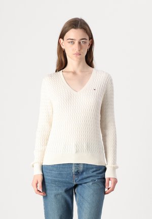 Pull crème en V avec un motif de torsades texturé, manches longues et ourlet à côtes. Petit logo sur la poitrine. Jeans visibles.