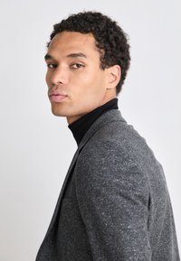 Blazer grigio con texture e un leggero sheen, indossato su un dolcevita nero. Il modello è di profilo, mettendo in evidenza la vestibilità sartoriale della giacca.