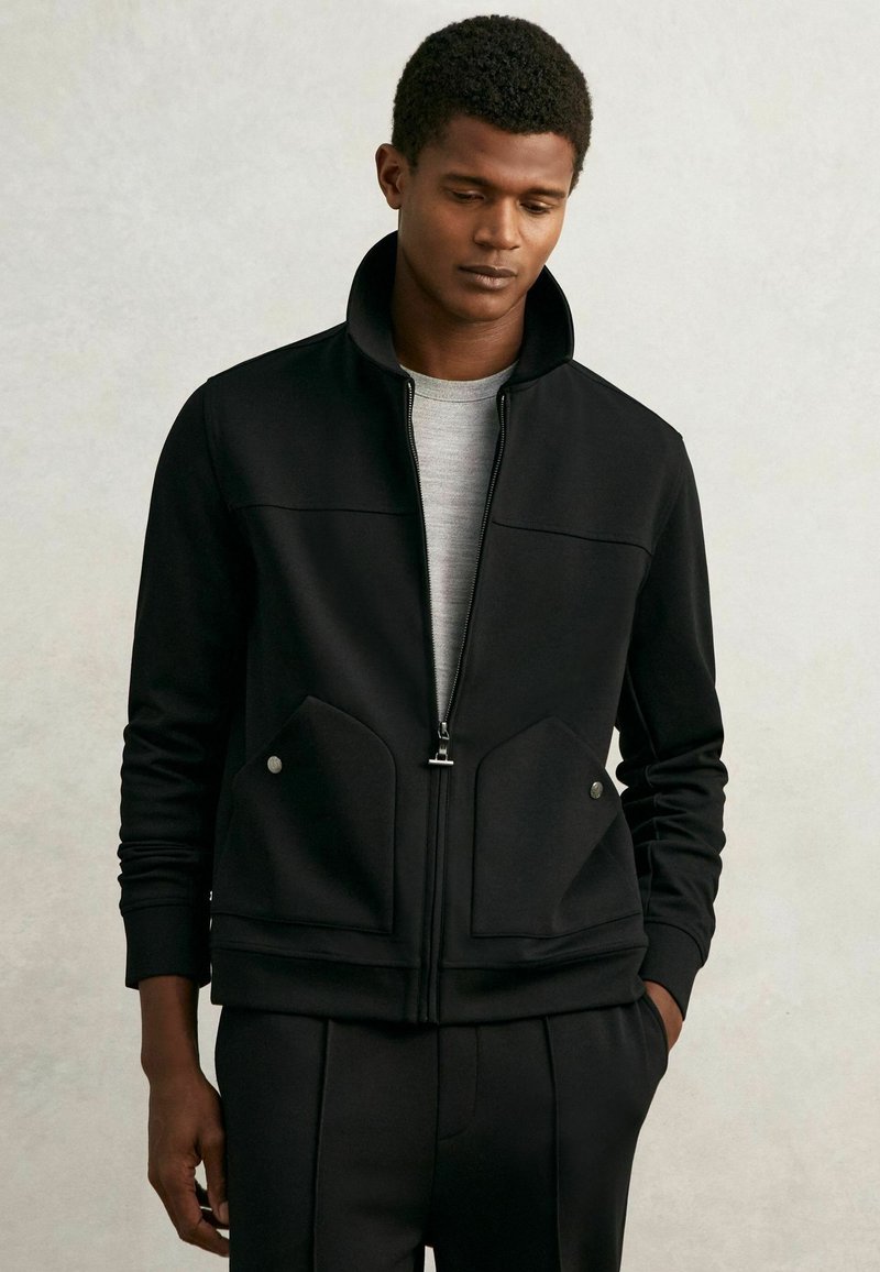 Schwarze Zip-Jacke mit hohem Kragen, ausgestattet mit zwei Fronttaschen und silbernem Zubehör. Hergestellt aus glattem, strukturiertem Stoff.