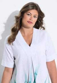 Weißes Leinenkleid mit kurzen Ärmeln, V-Ausschnitt und einem türkisfarbenen abstrakten Muster am Saum. Weiche Textur und lockere Passform.