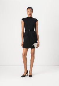 Robe noire à manches courtes avec un corsage ajusté, deux poches avant et une taille ceinturée. Associée à des escarpins noirs et une petite pochette blanche.