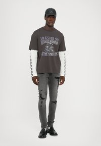Grijze graphic t-shirt met paarse print, gedragen over een witte lange mouwen shirt, gecombineerd met versleten grijze skinny jeans en zwarte schoenen.