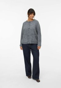 Cardigan gris texturizado con botones en el frente, dos bolsillos y cuello redondo, combinado con unos pantalones vaqueros de pierna ancha azul oscuro y zapatos marrones.