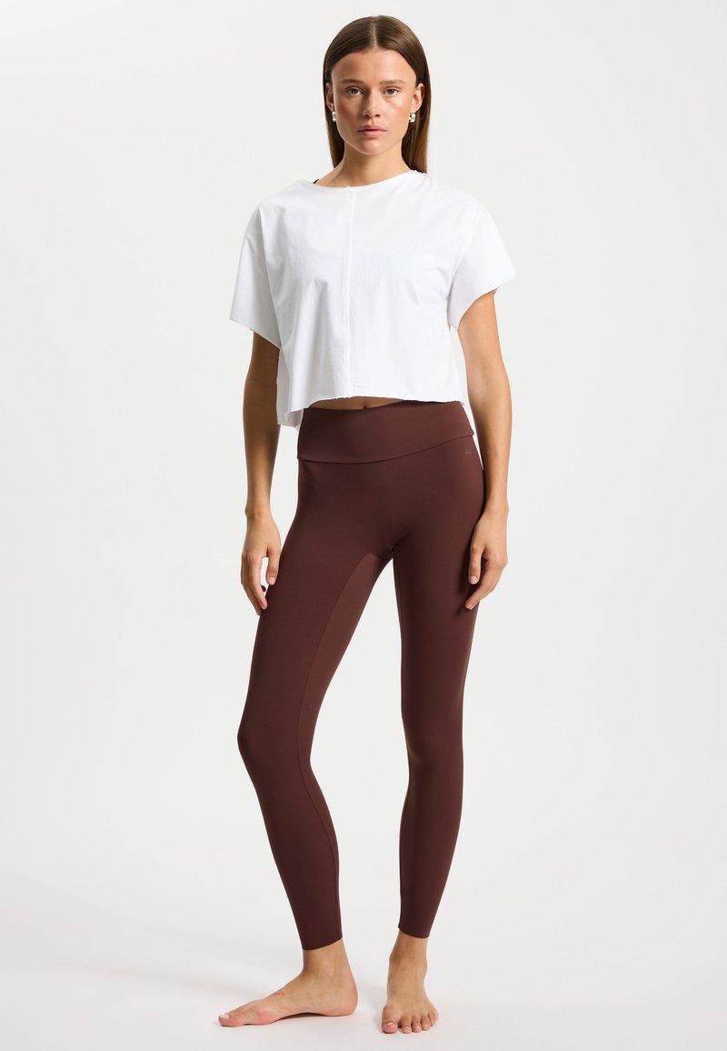 Witte cropped T-shirt met korte mouwen, gecombineerd met bruine, hoog getailleerde leggings. De leggings hebben een gladde afwerking en zijdetails.