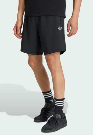 Mężczyzna ubrany w czarne spodenki Adidas, czarne skarpetki Adidas z białymi paskami oraz czarne buty sportowe Adidas, stojący na jasnym tle.