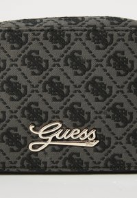 Svart texturerad tygplånbok med ett repetitivt logomönster och en framträdande gul "Guess"-logotyp som hårdvarudetalj.