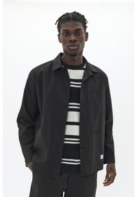 Black lightweight jacket with button closure, two front pockets, worn over a striped black and white sweater, against a white background.