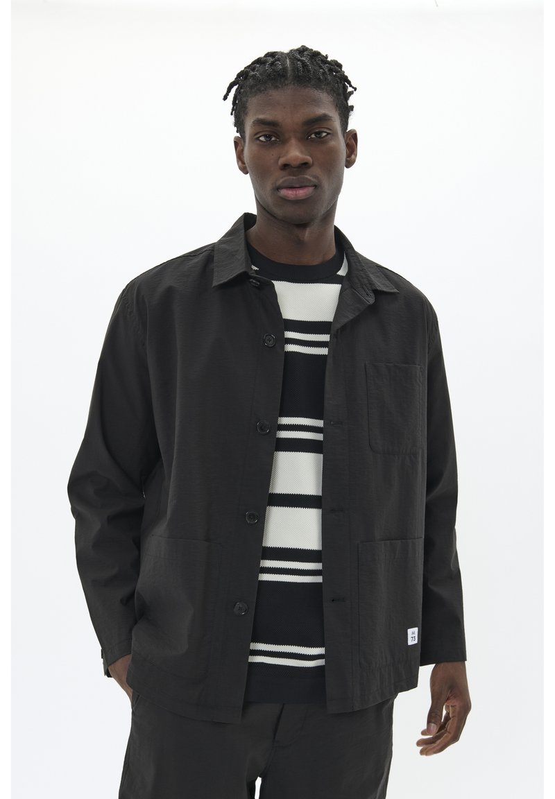 Black lightweight jacket with button closure, two front pockets, worn over a striped black and white sweater, against a white background.