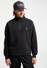 Felpa nera con zip e colletto alto, realizzata in tessuto morbido, con un piccolo logo bianco sul petto e polsini a coste.