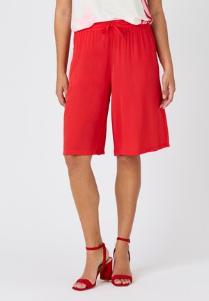 Donna che indossa pantaloncini rossi larghi fino al ginocchio con coulisse in vita e sandali rossi con tacco alto, in piedi davanti a uno sfondo semplice.