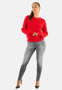 Donna in un maglione di lana rossa, jeans skinny grigi e tacchi alti neri, in piedi con le mani delicatamente incrociate, che guarda leggermente di lato.