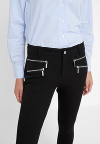 Pantalones negros con dos bolsillos con cremallera a cada lado. Hechos de un tejido suave. Combinados con una camisa de manga larga a rayas en azul claro.