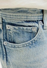 Jean en denim bleu clair avec une petite poche avant, des rivets en argent et des coutures visibles. La texture présente un motif classique délavé.