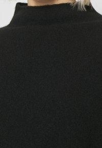 Pull noir en tissu duveteux à col haut, porté par une personne aux cheveux blonds clairs partiellement visibles, sur un fond uni.