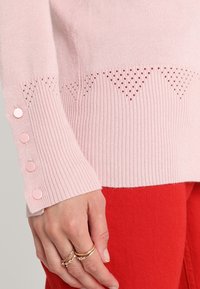 Różowy sweter dzianinowy o fakturze prążkowanej, z geometrycznym wzorem oczek i różowymi guzikami na rękawie.