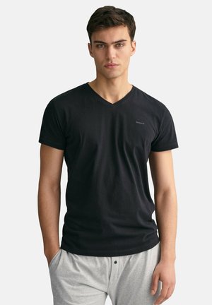 Schwarzes Baumwoll-T-Shirt mit V-Ausschnitt und kurzen Ärmeln, mit einem kleinen gestickten Logo auf der Brust. Kombiniert mit hellgrauen Hosen mit Kordelzug.