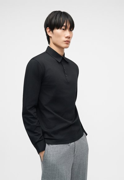 Άνδρας φορούντας μαύρο πόλο με μακριά μανίκια και γκρι παντελόνι με λεπτές ρίγες, με τα χέρια στις τσέπες, μπροστά σε απλό λευκό φόντο.