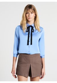 Blusa di raso blu chiaro con maniche lunghe, apertura con bottoni e dettaglio in cravatta nera al colletto. Abbinata a pantaloni corti a righe marroni.