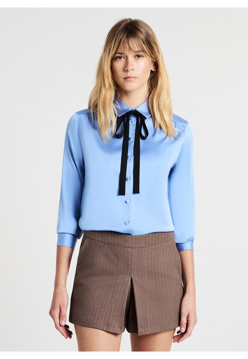 Blusa di raso blu chiaro con maniche lunghe, apertura con bottoni e dettaglio in cravatta nera al colletto. Abbinata a pantaloni corti a righe marroni.