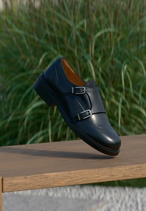 Zwarte leren schoen met dubbele monkstrap in balans op een houten bankje met een achtergrond van groen hoog gras.