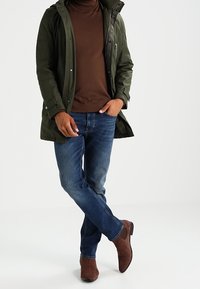 Mörkgrön vattentät parka med dragkedja och knäppning, brun polotröja, blå denimjeans och bruna ankelstövlar med en lätt klack.