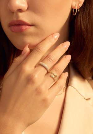 Gold- und Silberringe werden an den Fingern getragen. Die Hand ist in der Nähe des Gesichts positioniert. Die Nägel sind geformt und mit einer Nude-Farbe poliert.