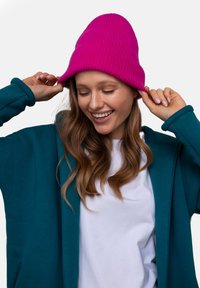 Turkusowa bluza z zamkiem na przodzie noszona na białej koszulce, w połączeniu z jaskrawym różowym, prążkowanym czapką. Długie faliste włosy i ręce poprawiające czapkę.