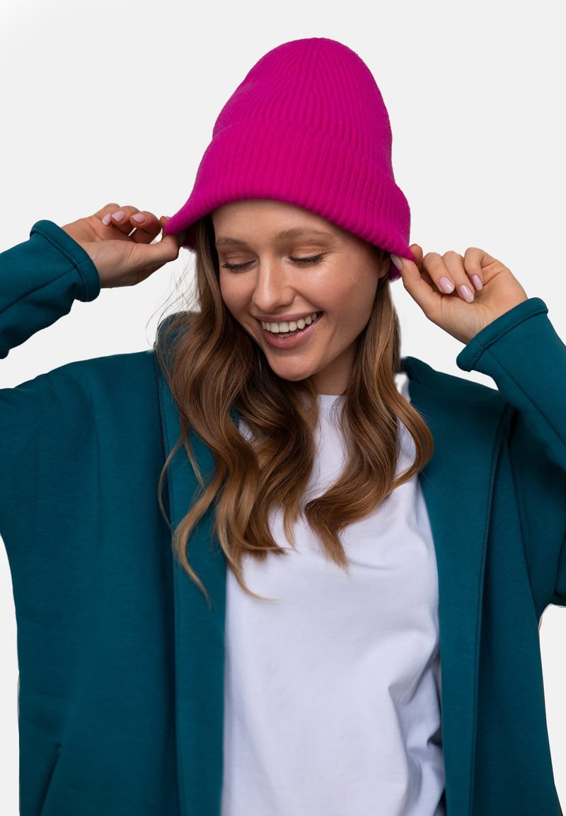 Turkusowa bluza z zamkiem na przodzie noszona na białej koszulce, w połączeniu z jaskrawym różowym, prążkowanym czapką. Długie faliste włosy i ręce poprawiające czapkę.