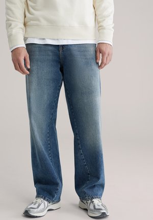 Lichtbeige sweatshirt over een wit T-shirt gedragen, gecombineerd met losse blauwe spijkerbroek met een vervaagde verloop en grijze sneakers.