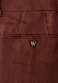 Gros plan sur un tissu marron avec une poche passepoilée boutonnée, montrant les détails de la couture et la texture du matériau.