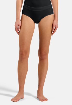 Femme portant une culotte noire sans coutures taille haute, debout pieds nus sur un fond blanc uni.