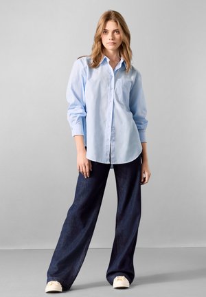 Helles blaues Hemd mit langen Ärmeln und Brusttasche, kombiniert mit dunklen, weit geschnittenen Jeans und beigen Schuhen.