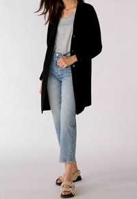 Svart lång kappa, ljusgrå V-ringad topp, urblekta blå jeans med hög midja, beige texturerade sandaler med två remmar och en tjock sula.