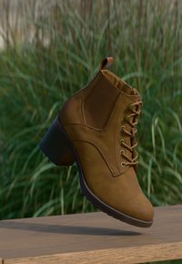 Braune Leder-Ankle-Boots mit Blockabsatz und Schnürung, positioniert am Rand einer Holzfläche mit hohem Gras im Hintergrund.