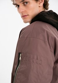 Brun bomberjacka med en ficka med dragkedja och en texturerad svart huva. Slät tyg med en något vadderad struktur.