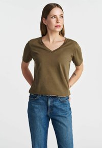 Olivewnozielona koszulka z dekoltem w serek, wykonana z gładkiego materiału, z krótkim rękawem, w luźnym kroju, zestawiona z niebieskimi dżinsami. Prosty design, bez wyraźnych wzorów.