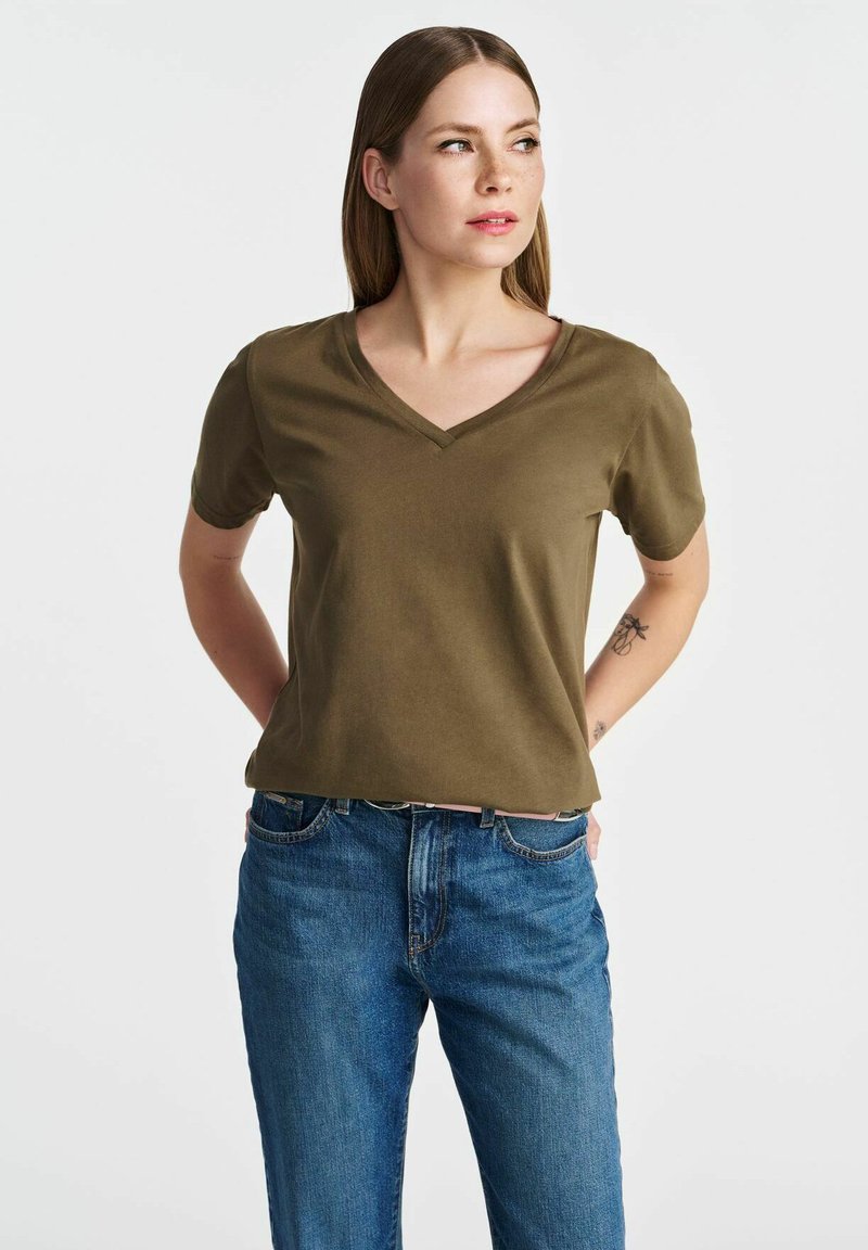 Olivewnozielona koszulka z dekoltem w serek, wykonana z gładkiego materiału, z krótkim rękawem, w luźnym kroju, zestawiona z niebieskimi dżinsami. Prosty design, bez wyraźnych wzorów.